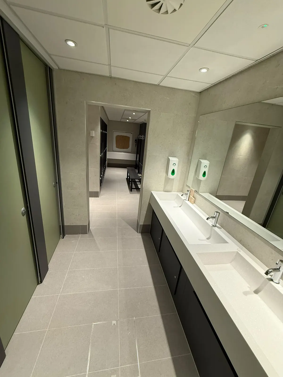 Washroom at All Seasons Leisure Centre in Chorley, with twin basins on a dark vanity run below a full-width mirror and Unika Light wall tiles, looking through to the changing room