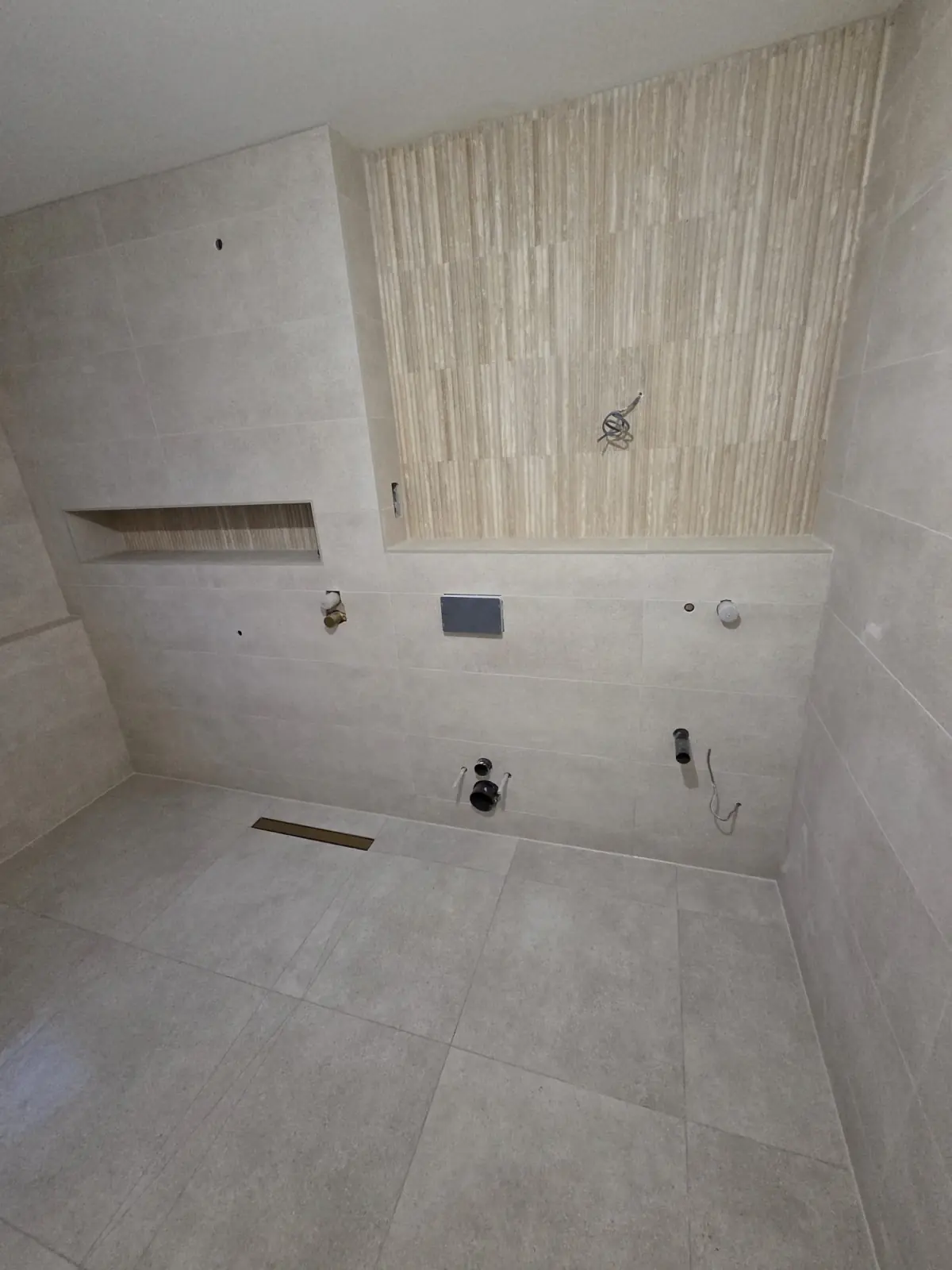 Wet room before fit-out with a fluted travertine-effect feature wall, tiled niche and brushed brass linear drain