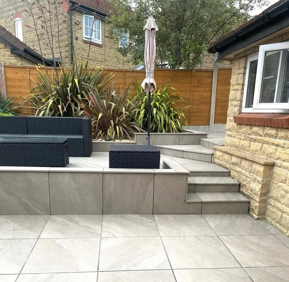 Light grey outdoor porcelain patio with a raised seating area, tiled planters and steps beside a sandstone wall